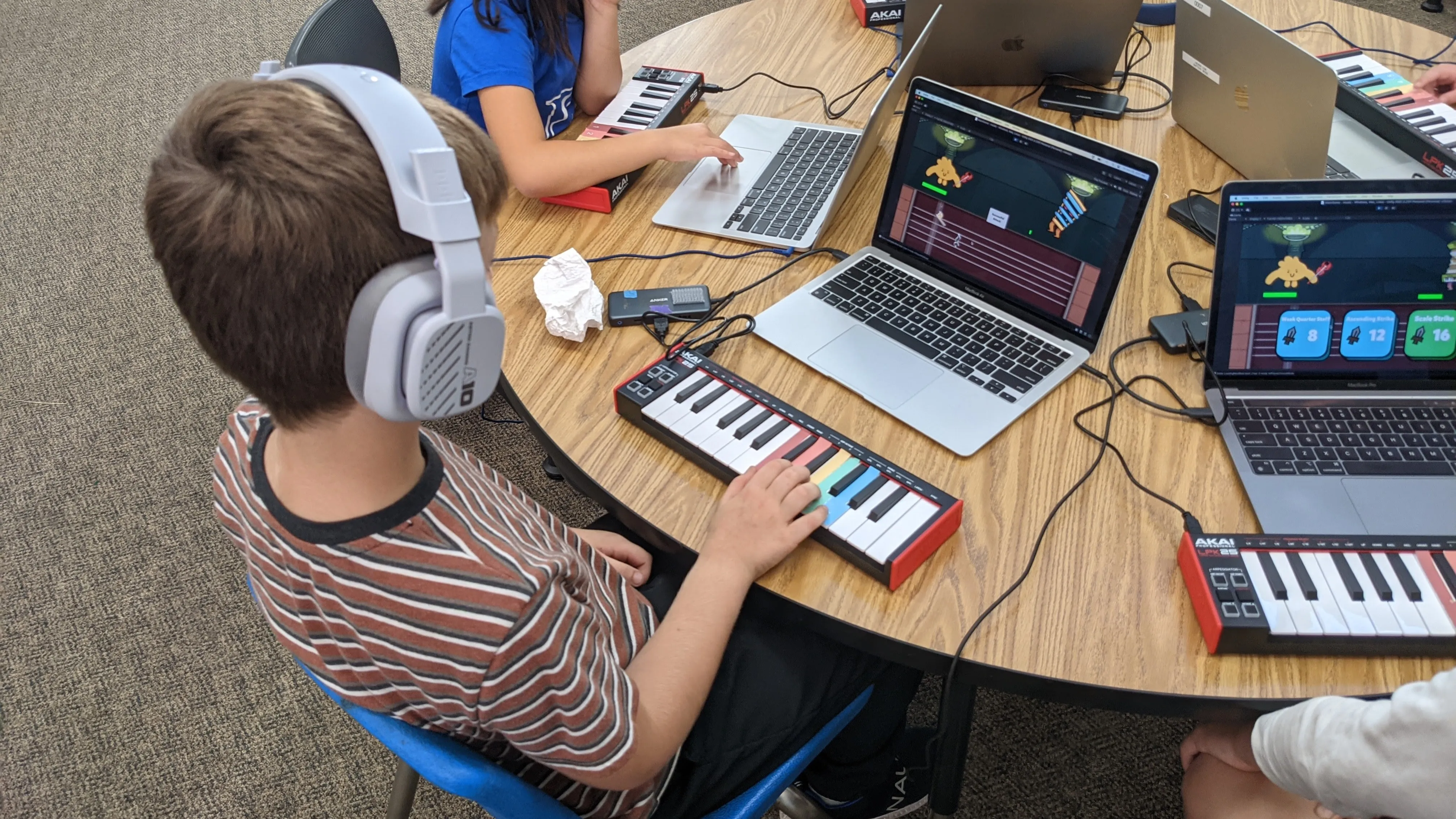Students around a shared table playing the game on laptops, each with a MIDI keyboard in front of them; the nearest wears headphones, one hand on the keys.