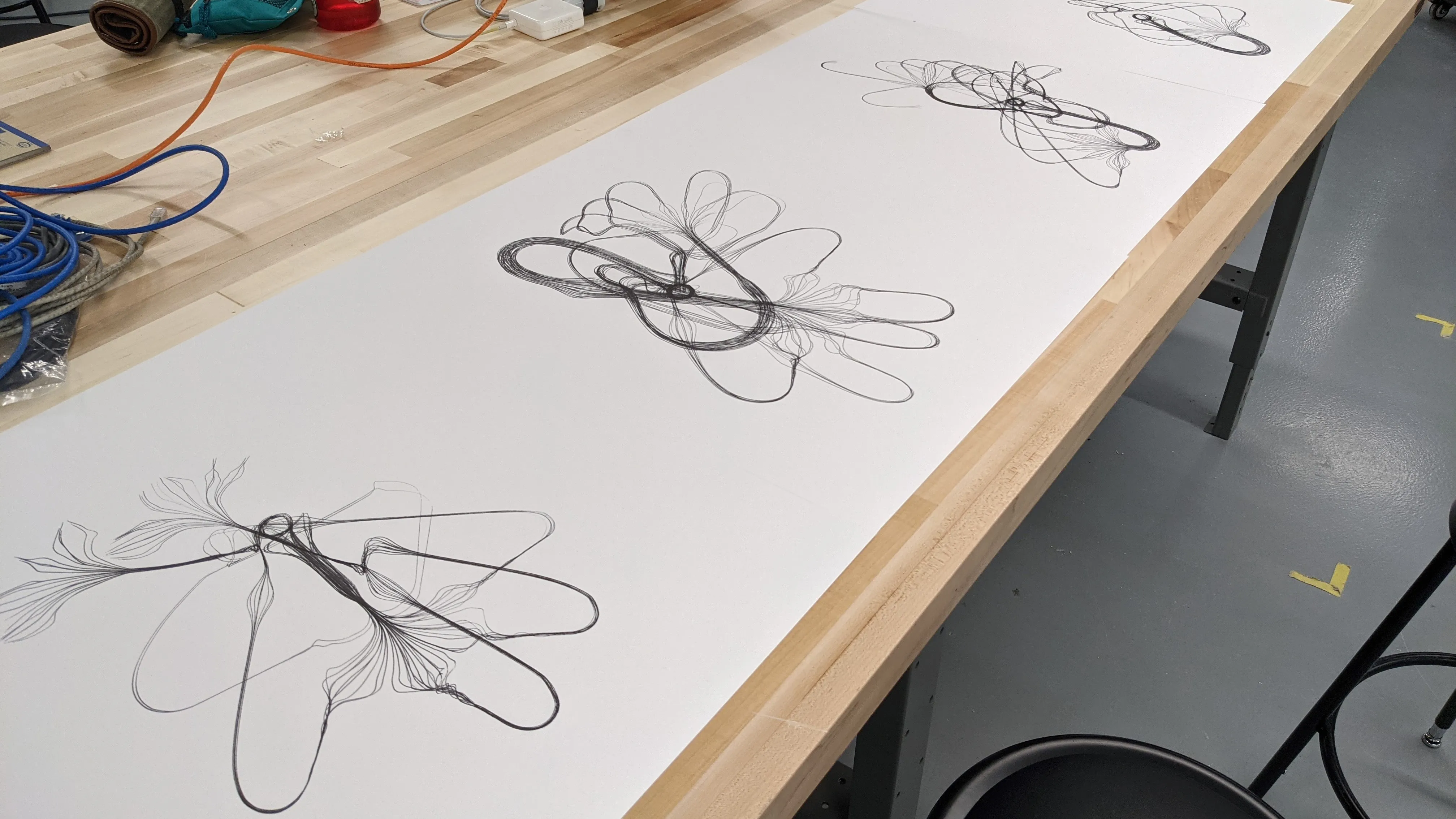 A wide roll of paper along a workbench, plotted with three large looping drawings, each built from a single continuous line.